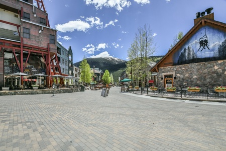 A spacious outdoor plaza with brick paving framed by rustic buildings, trees with budding leaves, outdoor seating, and a snow-capped mountain in the background under a clear blue sky.