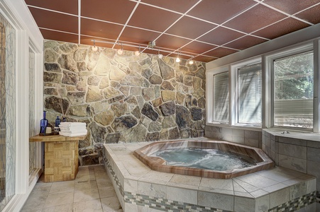 Cozy bathroom featuring a stone wall, a large hot tub with beige tiles, a small wooden table with towels, and ample natural light coming through adjacent windows.