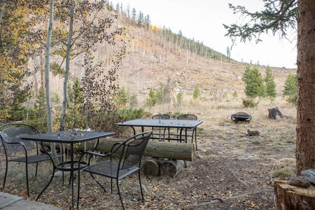 Outdoor area with metal tables and chairs set in a grassy, partially forested landscape with a hill in the background. A fire pit is visible on the right side of the image.