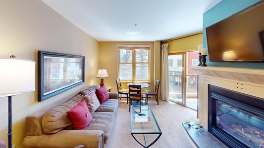 A cozy living area with a brown sofa, a glass coffee table, a round dining table with two chairs by a window, and a mounted flat-screen TV above a fireplace. Natural light illuminates the room.