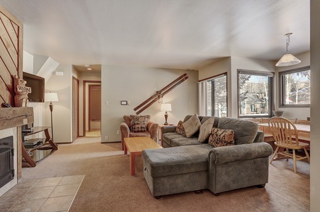 A cozy living area with a green sectional sofa, wooden coffee table, a fireplace, wall decor including skis, and a dining table in front of windows showing an outdoor view.