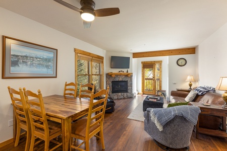 A cozy living and dining area featuring a wooden dining table with chairs, a lounge with a stone fireplace, a flat-screen TV, and a balcony door. Warm lighting and a ceiling fan are also visible.