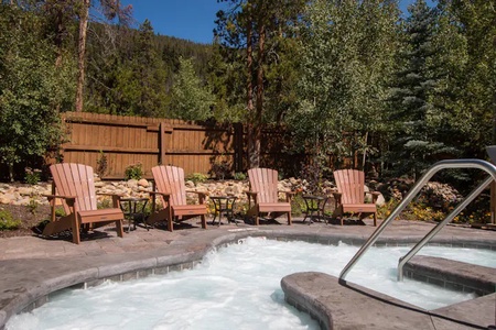 A bubbling hot tub surrounded by wooden lounge chairs and tables is set against a backdrop of trees and a wooden fence.