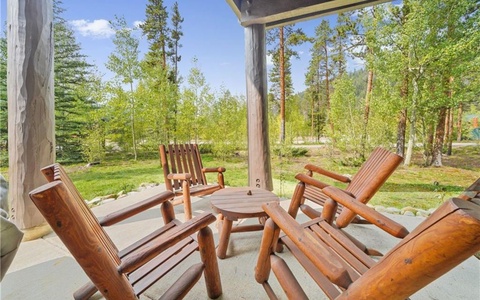 Wooden patio furniture, including four chairs and a round table, is arranged on a concrete porch, overlooking a green wooded area on a sunny day.