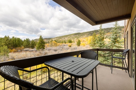 A balcony with a black metal table and chairs overlooks a scenic landscape of trees and hills under a cloudy sky.