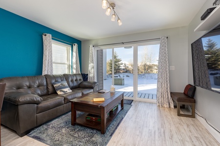 A modern living room with a gray sofa, wooden coffee table, and a wall-mounted TV. Large sliding glass doors open to a deck with a snowy view outside. The left wall is painted teal.