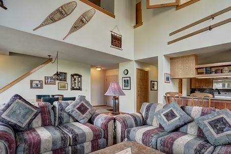 A living room with patterned sofas, a wooden coffee table, and decor including snowshoes and photo frames. An open kitchen with wooden cabinets is visible in the background.