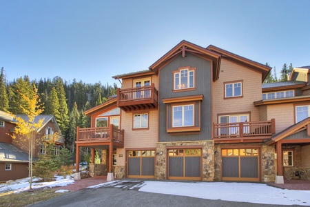 A multi-story house with wooden balconies, multiple garages, and a stone facade set against a backdrop of pine trees and a clear sky. Snow is scattered on the driveway and surrounding ground.