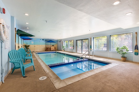 An indoor swimming pool with deck chairs, potted plants, and large windows providing natural light. There is a small table with chairs and umbrellas visible in the background.