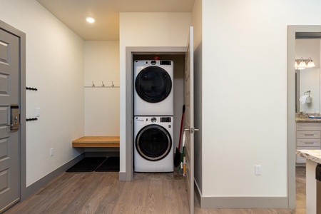 Laundry room with a stacked washer and dryer unit in a closet, adjacent to a wooden bench with hooks on the wall, and a door leading to a bathroom.