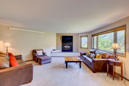 A cozy living room with two brown leather sofas, an armchair, a wooden coffee table, lamps, a fireplace, and a wall-mounted TV. Large windows provide natural light and a view of greenery outside.