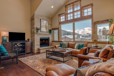 A cozy living room features leather sofas, a large rug, a wooden coffee table, a fireplace with a stone surround, and a flat-screen TV. Large windows offer a view of snowy rooftops and trees outside.