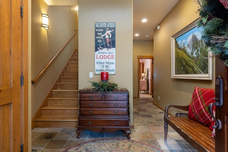 Warm and inviting hallway with a wooden staircase, antique chest of drawers, wall art, and a bench adorned with a red plaid cushion. Wall sconce lighting and evergreen wreath create a cozy atmosphere.