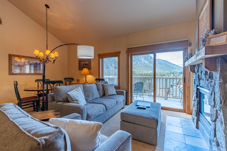 A living room with two gray sofas, a large window with mountain views, a fireplace, dining area, and a sliding glass door opening to a balcony with outdoor seating.