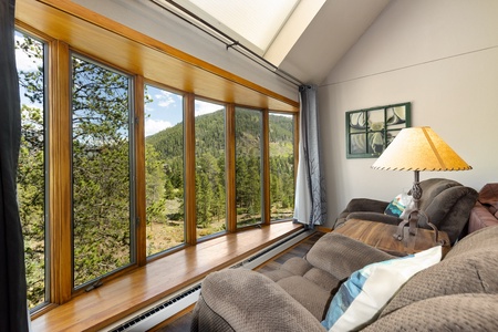 A cozy living room with large windows offering a scenic view of a lush, forested mountain landscape. The room features a comfortable sofa, a wooden table, and a lamp.