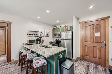 A modern kitchen with a granite island, colorful bar stools, stainless steel appliances, wooden cabinets, and a wooden door. The room features white walls and a hardwood floor.