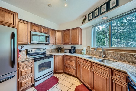 A modern kitchen with wooden cabinets, stainless steel appliances, granite countertops, a double sink, and a large window overlooking trees. Decor includes framed artwork and a potted plant.