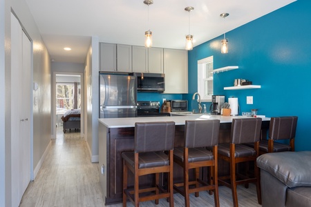 Modern kitchen with blue accent wall, gray cabinets, stainless steel appliances, and white countertops. There is a large island with four brown chairs and pendant lighting hanging above.