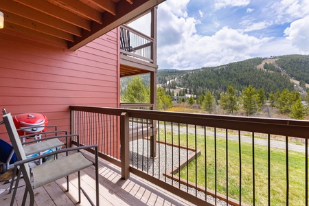 A wooden deck with two chairs and a barbecue grill overlooks a grassy area and tree-covered hills under a partly cloudy sky.