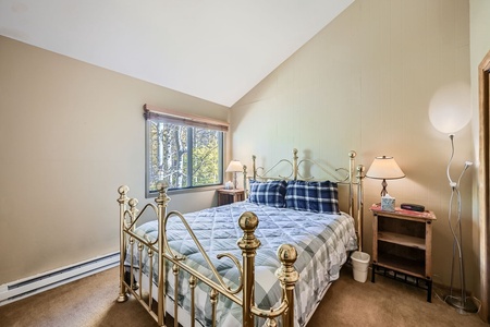 Bedroom with a brass bed, plaid bedding, two bedside tables with lamps, a window with a view of trees, and beige walls.