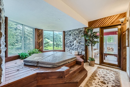 A room with a large hot tub on a wooden platform, surrounded by windows showing a forested view, and a door with glass panels. The room has stone walls and various potted plants.