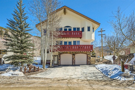 A three-story house with white walls and red balconies, set in a snowy landscape with pine trees and mountains in the background. The house has a two-car garage and a driveway.