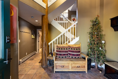 A small foyer with a wooden bench, decorative pillows, a small Christmas tree, and a wooden column. A staircase with white railings is in the background, and a hallway leads to other rooms.