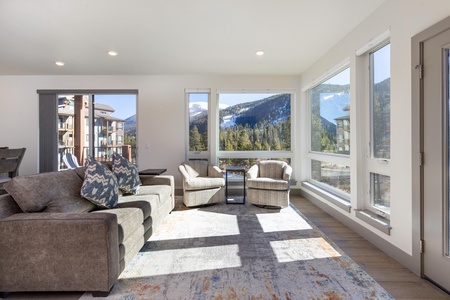 Modern living room with large windows offering a mountain view. Room features a sofa, two armchairs, a coffee table, and a patterned rug. Natural light floods the space.