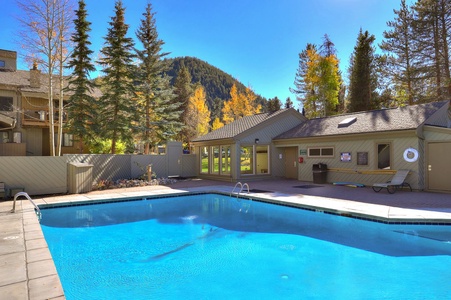 Outdoor pool area on a sunny day with a view of trees and a mountain in the background. The pool is surrounded by a concrete patio, a fence, and wooden buildings.