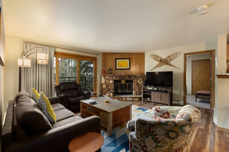 A cozy living room with a stone fireplace, TV, assorted seating, and wooden decor. Large window and sliding door on the left provide natural light. A mixture of modern and rustic furnishings.