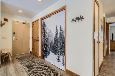 A hallway with wooden doors, a black and white snow-covered forest mural, a wooden bench, a patterned rug, and a wall-mounted coat rack with animal figures. The floor seems wooden.