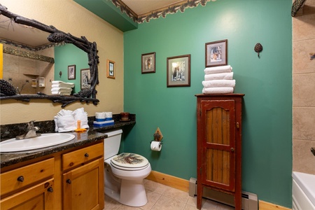 A bathroom with green and beige walls, wooden cabinets, a sink, toilet, a red wooden cabinet, framed pictures, and neatly folded towels on the counter and the red cabinet.