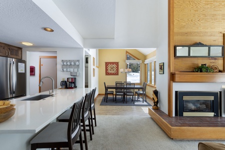 A modern open-concept kitchen with a white countertop adjacent to a dining area, featuring a wooden dining table and chairs. The room includes large windows and a fireplace with a wooden mantel.