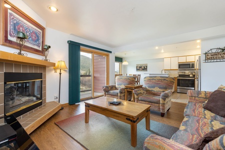 A living room with a fireplace, wooden coffee table, patterned chairs, and a sliding glass door leading to a balcony. The kitchen in the background has stainless steel appliances and a "Winter Welcome" sign.