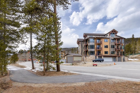 A modern multi-story apartment building with balconies set against a mountainous backdrop. There are pine trees, a paved path, and a parked vehicle near the building. The sky is partly cloudy.