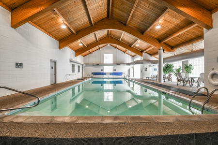 An indoor swimming pool with a wooden ceiling, lounge chairs, potted plants, and pool rules posted on the wall. The area is well-lit with large windows and overhead lights.