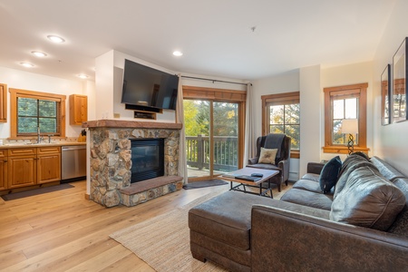 A cozy living room with a stone fireplace, mounted TV, sectional sofa, coffee table, and large windows. The kitchen with wooden cabinets is visible in the background.