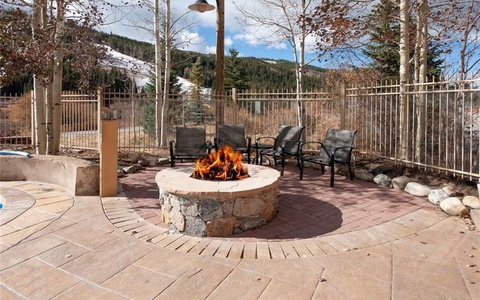 Outdoor patio with a stone fire pit, surrounded by four metal chairs, set against a background of trees and a fenced area. Snow-covered hills are visible in the distance under a partly cloudy sky.