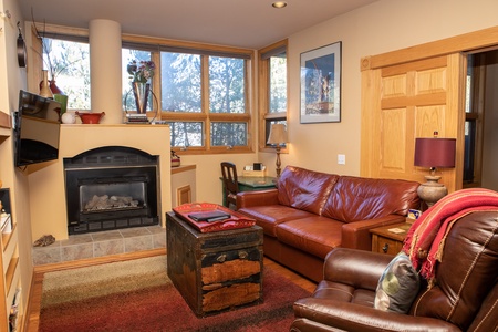 A cozy living room with a brown leather couch, a fireplace, wooden furniture, and a large window letting in natural light. A colorful rug covers the floor, and a rustic chest serves as a coffee table.