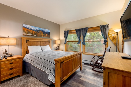 A neatly arranged bedroom featuring a wooden bed frame with a striped comforter, nightstands with lamps, a wall-mounted TV, and a wooden desk with a chair. Large windows offer a view outside.