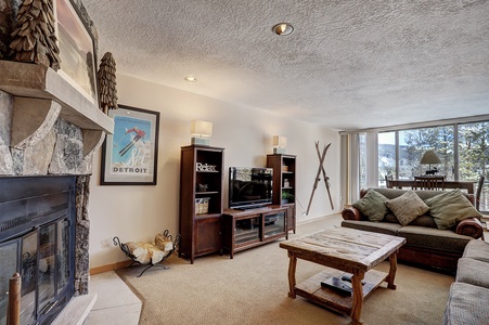 A cozy living room with a stone fireplace, wood furniture, a TV, skis on the wall, and a large window offering a scenic view. A framed "Detroit" poster is displayed above the fireplace.