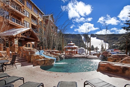 Outdoor pool area with a waterslide, lounge chairs, and a gazebo, surrounded by a hotel building. Snow-covered mountains and blue sky are visible in the background.