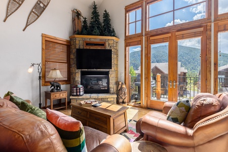 Cozy living room with a fireplace, large windows showcasing a mountain view, and rustic decor including snowshoes and wooden furniture.