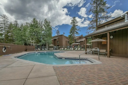 An outdoor swimming pool with lounge chairs and umbrellas, surrounded by trees and wooden buildings under a partly cloudy sky.