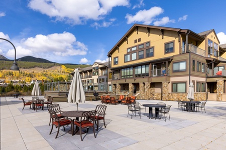 Outdoor patio area with tables, chairs, and umbrellas in front of a large building on a sunny day. The backdrop features mountains and trees with fall foliage under a blue sky with scattered clouds.