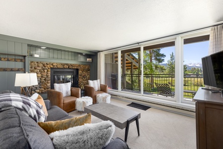 A cozy living room features a stone fireplace, gray and brown furniture, and large windows overlooking a lush outdoor landscape with trees and mountains.