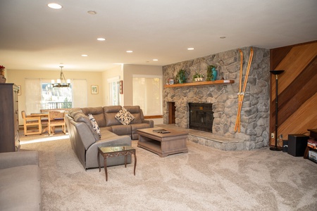 A cozy living room with a stone fireplace, a sectional sofa, a coffee table, and carpeted flooring. A dining area is visible in the background with a wooden table and chairs. Decor includes wooden panels and skis.