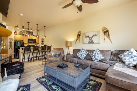 Cozy living room with a large sectional sofa, coffee table, and wall-mounted TV. Kitchen area with barstools visible in the background. Warm, neutral decor and snowshoes hanging on the wall.