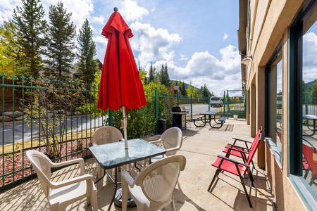 Outdoor patio with a glass table, several white chairs, two red chairs, and a red umbrella. Green fence and trees in the background with a partly cloudy sky.