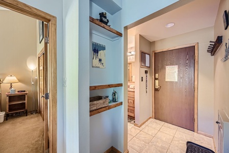 A hallway with a wooden door, built-in shelves, and tile flooring. A room with a lamp and furniture is visible to the left.